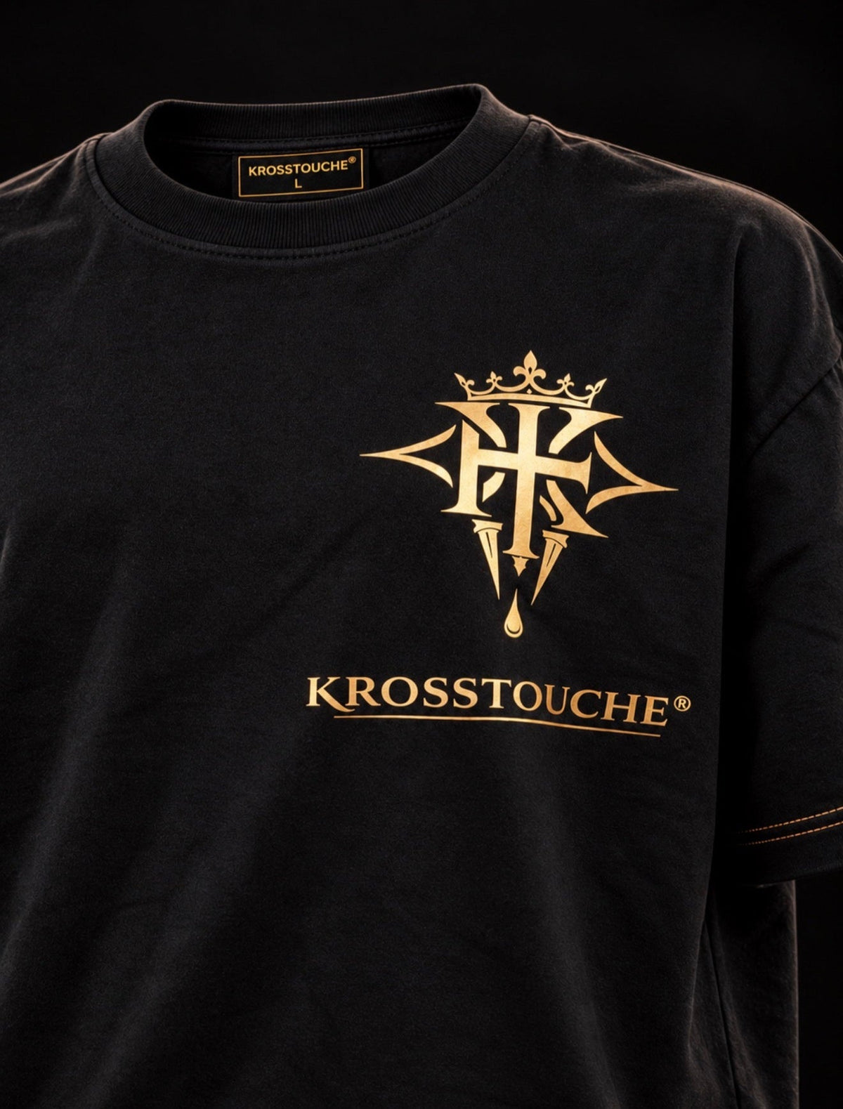Black 300 GSM oversized KROSSTOUCHE® T-shirt shown from a three-quarter front view, featuring the original gold crown and cross emblem with the KROSSTOUCHE® wordmark beneath it, gold double stitching on the sleeve cuffs, against a dark studio background.