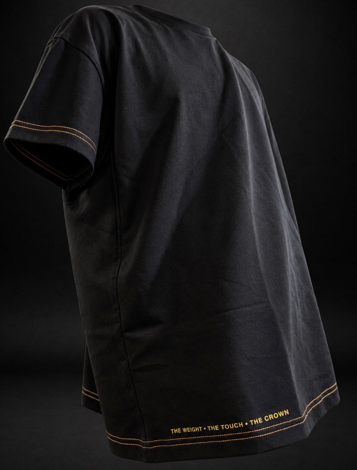 Side view of a black KROSSTOUCHE® luxury T-shirt showcasing its oversized silhouette, premium gold double stitching along the sleeve and bottom hem, and the signature gold lettering THE WEIGHT • THE TOUCH • THE CROWN near the lower hem.