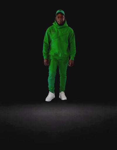 Model wearing the KROSSTOUCHE® Ascension emerald green hoodie, panelled joggers and matching cap against a black studio background.